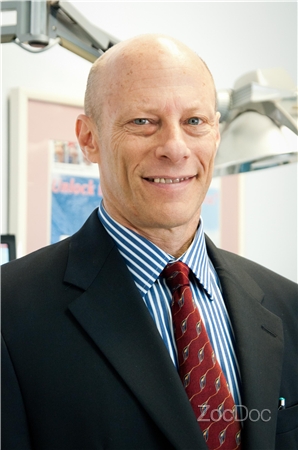 The image shows a man standing in front of a background that includes a sign with the word  ZOODOC  and what appears to be medical equipment, suggesting he may be associated with a veterinary or animal healthcare facility. He is wearing a suit and has a bald head, smiling slightly at the camera.