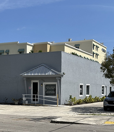 The image shows a one-story building with a white facade and a flat roof, featuring a prominent entrance with a small overhang. There is a sign that reads  Hampton Inn  above the door, and the sky is clear with no visible clouds.