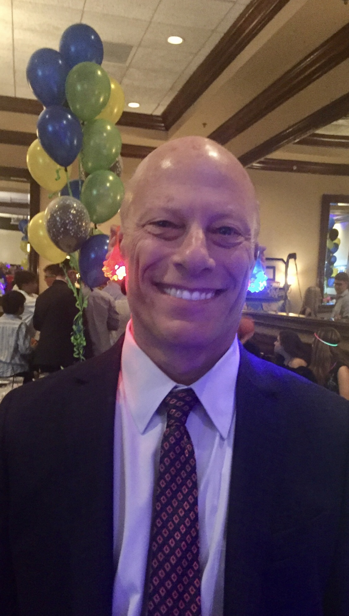 The image is a photograph of a man standing in front of a balloon arch at an indoor event, smiling and looking directly at the camera.
