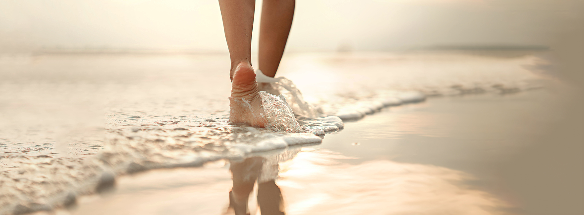 A person is walking along a sandy beach towards the water, with their feet creating ripples in the shallow waves.